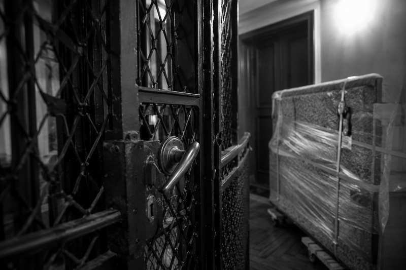 Piano droit emballé en attente sur un palier devant une cage d’ascenseur en fer forgé trop étroite