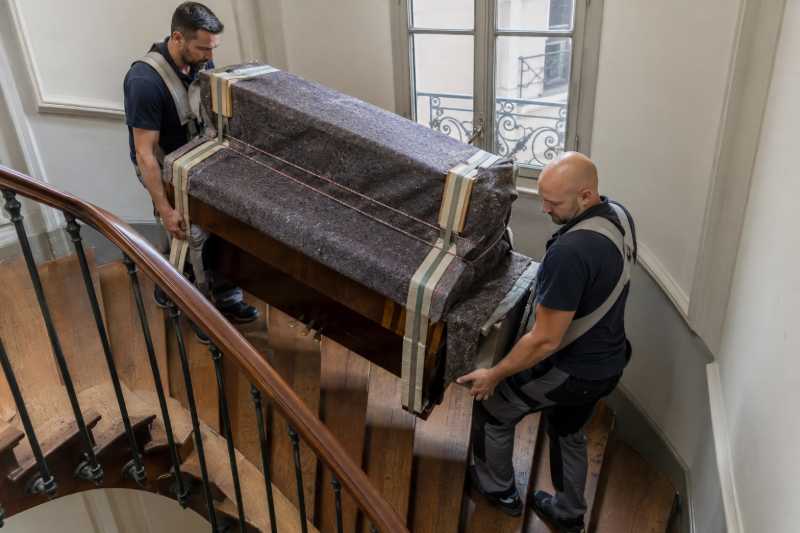Deux déménageurs au harnais descendant un piano droit emballé dans un escalier d'immeuble, Antony