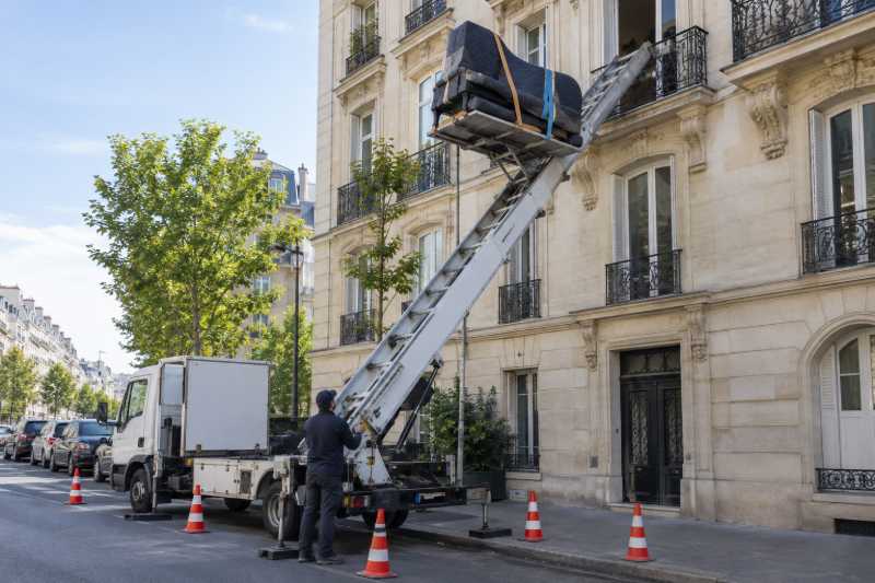 Camion monte-meubles hissant un piano à queue emballé le long d'une façade en pierre, Orsay